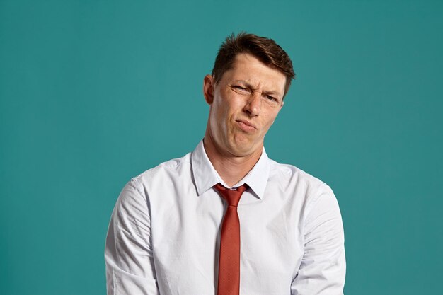 Studio portrait of a funny young businessman in a classic white shirt and red tie looking disappointed while posing over a blue background. Stylish haircut. Sincere emotions concept. Copy space.