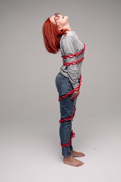 鎖に絡まった少女のスタジオポートレート茶髪の赤髪の女性が鎖を取り除こうとするリアルな状況のコンセプト灰色の背景に分離