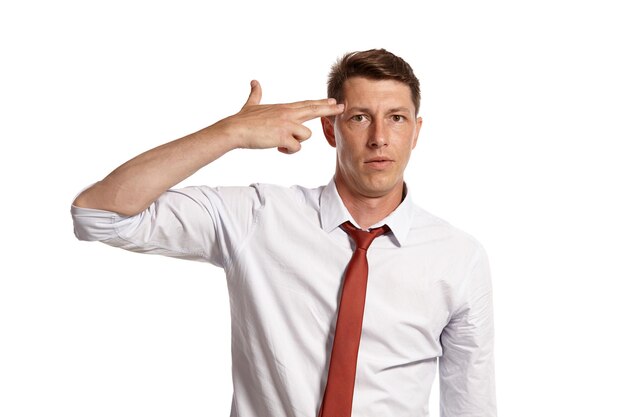 Studio portrait of a good-looking young man in a classic white shirt and red tie shooting himself from a finger while posing isolated on white background. Stylish haircut. Sincere emotions concept. Co