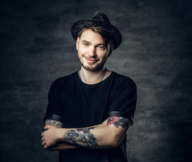 Studio portrait of handsome male with crossed tattooed arms, dressed in a black t shirt and tweed hat.