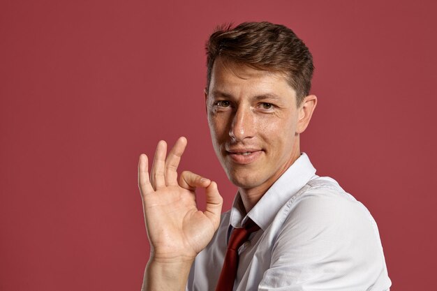 Studio portrait of a handsome young man in a classic white shirt and red tie, smiling and showing ok sign while posing over a pink background. Stylish haircut. Sincere emotions concept. Copy space. Cl