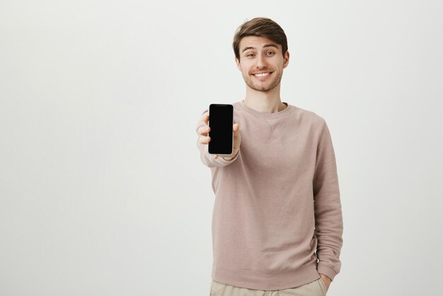 笑顔でポケットに片手を持ってスマートフォンを示す幸せな魅力的なヨーロッパの男性モデルのスタジオポートレート