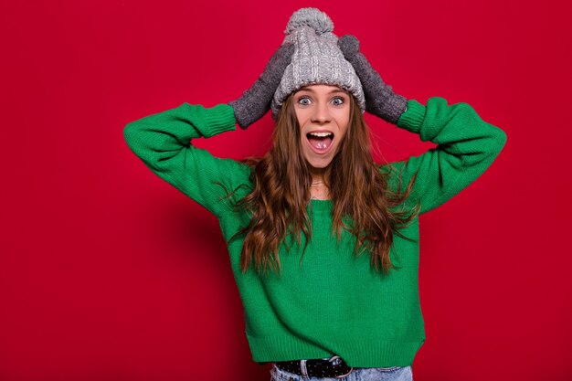 Studio portrait of lovely exited happy woman with long light-brown hair wearing green pullover and grey winter cap posing in the camera with open mouth and holds up hands, isolated background