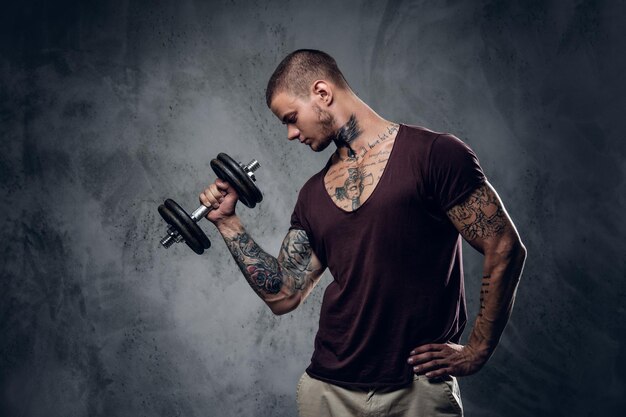 彼の腕に入れ墨をしたシャツを着た男のスタジオポートレートは、灰色の芸術的な背景の上にダンベルを保持しています。