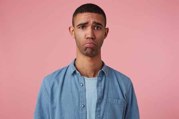 Ritratto in studio di mezza età uomo mediorientale indossa camicia blu guarda nella telecamera con espressione facciale triste, curvando il labbro isolato su sfondo rosa