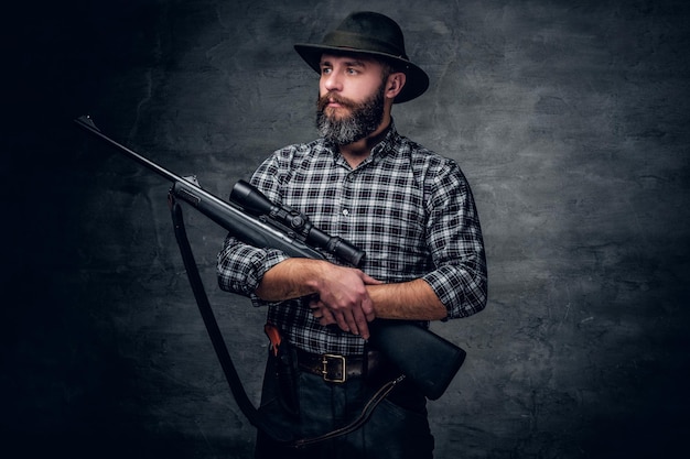 Бесплатное фото Студийный портрет бородатого охотника в клетчатой флисовой рубашке с винтовкой.