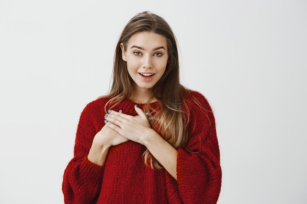 Studio portrait of pleased flattered young attractive woman in red loose sweater, holding palms on chest and smiling broadly, speaking with boyfriend who confessing in love 