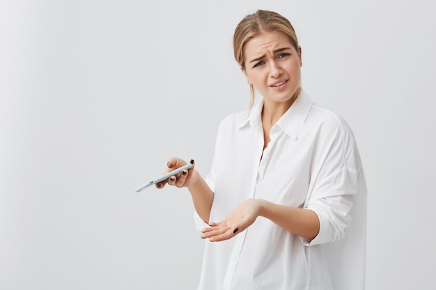 Studio portrait of puzzled or anxious pretty girl with blonde hair frowning her face looking at phone seeing bad news or photos posing . Negative human emotion, reaction.