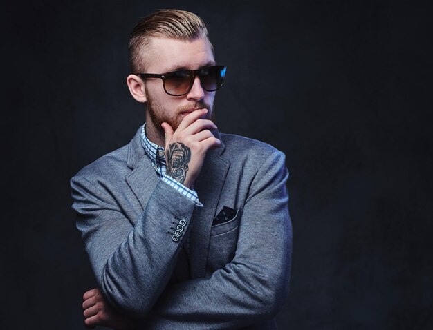 Studio portrait of redhead bearded male dressed in a suit and sunglasses over grey background.