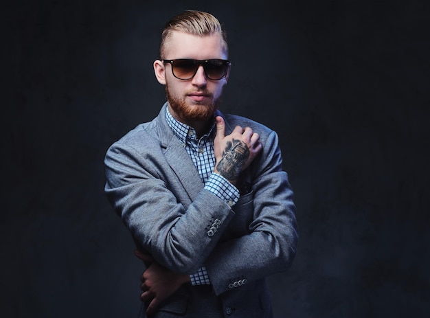 Studio portrait of redhead bearded male dressed in a suit and sunglasses over grey background.