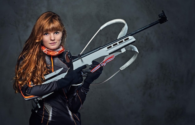 Studio portrait of a redhead female Biathlon champion holds a gun over grey background.
