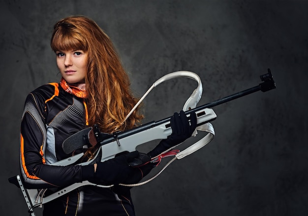 Studio portrait of a redhead female Biathlon champion holds a gun over grey background.