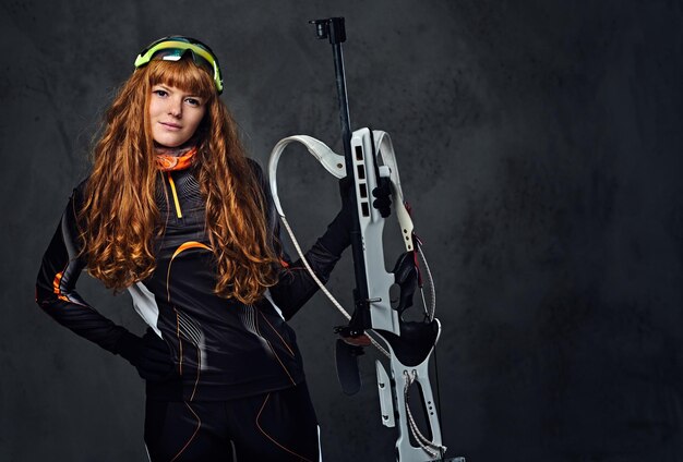 Studio portrait of a redhead female Biathlon champion holds a gun over grey background.