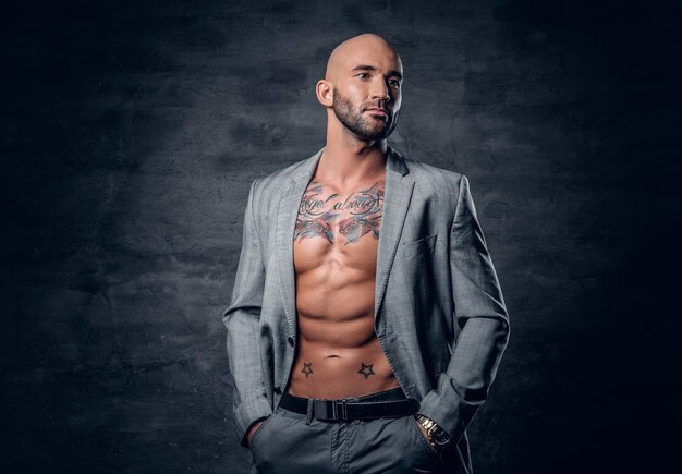 Studio portrait of shaved head tattooed male dressed in a grey jacket on a naked torso.