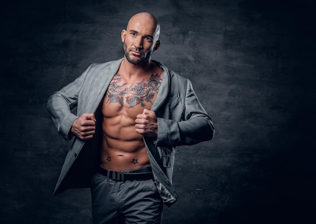 Studio portrait of shaved head tattooed male dressed in a grey jacket on a naked torso.