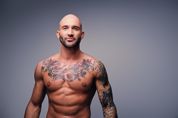 Studio portrait of shirtless athletic shaved head male with tattoos on his torso posing over grey vignette background.
