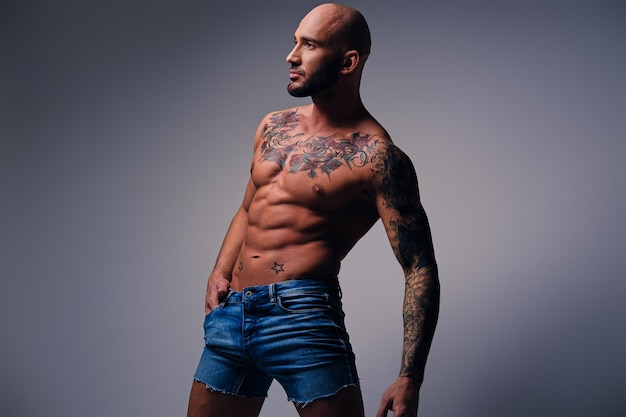 Studio portrait of shirtless shaved head, muscular male with tattoos on his torso dressed in a denim shorts over grey vignette background.