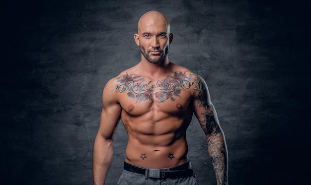 Studio portrait of shirtless shaved head, muscular male with tattoos on his torso over grey vignette background.