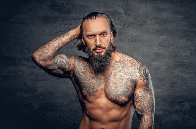 Studio portrait of shirtless, tattooed bearded male over dark grey vignette background.