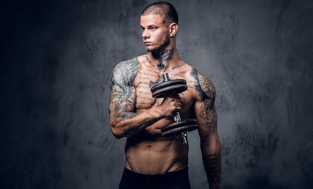 Free photo studio portrait of shirtless, tattooed male holds dumbbell over grey artistic background.