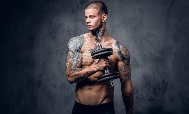 Studio portrait of shirtless, tattooed male holds dumbbell over grey artistic background.