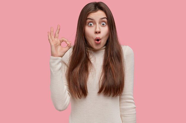 Studio portrait of shocked amazed woman makes okay gesture with suprised expression, stands over pink background, dressed in white casual jumper