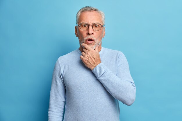 Studio portrait of shocked grey haired senior man holds chin keeps mouth opened hears something astonishing wears long sleeved jumper isolated over blue wall