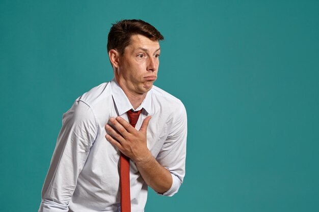 Studio portrait of a smart young male in a classic white shirt and red tie standing sideways and acting like he is sick while posing over a blue background. Stylish haircut. Sincere emotions concept.