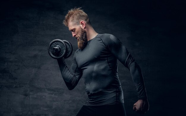 Studio portrait of sporty bearded male dressed in a grey sportswear holds dumbbell.