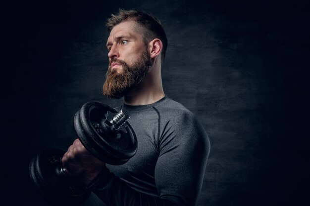 Studio portrait of sporty bearded male dressed in a grey sportswear holds dumbbell.