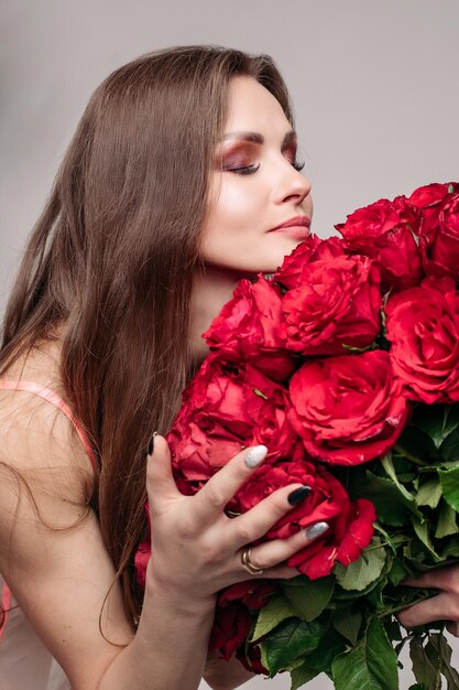 目を閉じて赤いバラの大きな束の匂いを嗅ぐセクシーなナイトドレスを着てメイクアップで見事な若いブルネットの女性のスタジオポートレートバラの香りを楽しんでいます