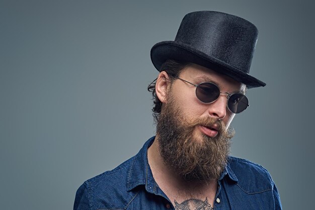 Studio portrait of stylish bearded male with a tattoo on his chest, dressed in a denim shirt, cylinder hat and sunglasses isolated on grey background.