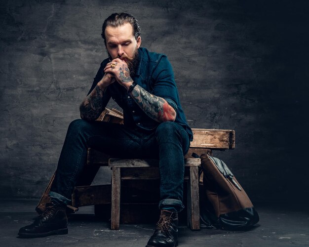 Студийный портрет стильного бородатого мужчины с татуировками на руках, сидящего на деревянных ящиках на сером фоне в студии.