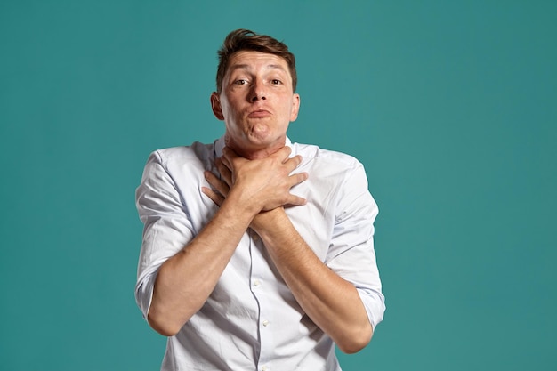 Free photo studio portrait of a stylish young male in a classic white shirt choking himself and looking at the camera while posing over a blue background. stylish haircut. sincere emotions concept. copy space.