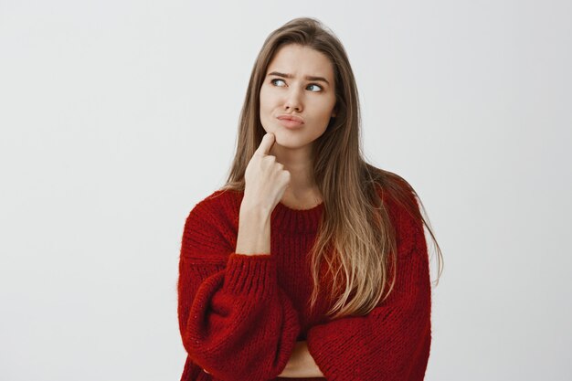 Studio portrait of thinking attractive european woman, feeling confused and troubled, looking up with concerned expression, touching chin with index finger, looking for solution 