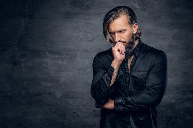 Studio portrait of thoughtful bearded male with long hair dressed in black jacket on grey background.