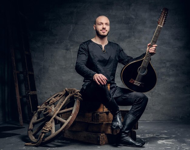 Studio portrait of traditional folk musician dressed in vintage Celtic clothes sits on a wooden box and holds mandolin.