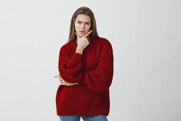 Studio portrait of troubled doubtful attractive woman in stylish red loose sweater, holding gun gesture on chin and frowning, feeling suspicious and frustrated, standing 