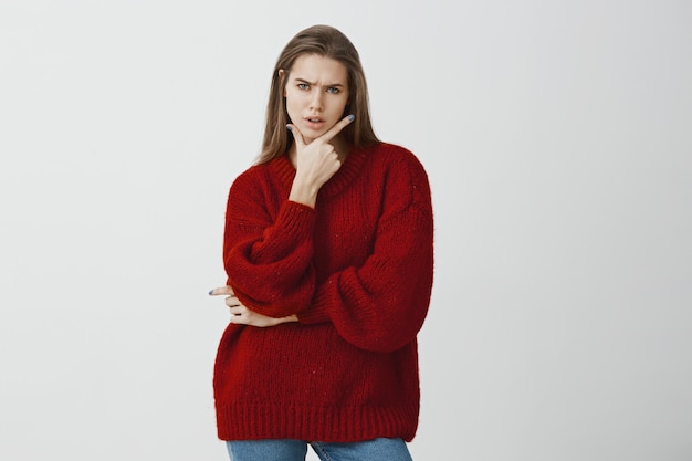 Студийный портрет беспокойной сомнительной привлекательной женщины в стильном красном свободном свитере, держа пистолет жестом на подбородке и нахмурившись, чувствуя подозрение и разочарование, стоя