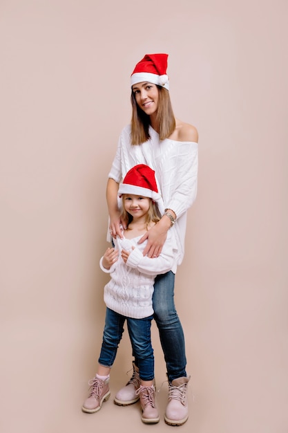 Studio portrait of young attractive woman with her little daugther wearing white sweaters and Christmas hats