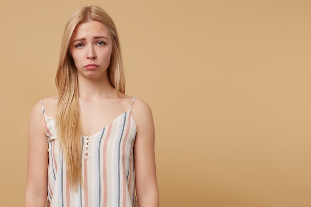 Studio portrait of young blonde woman posing over beige background curving her lip with sad facial expression
