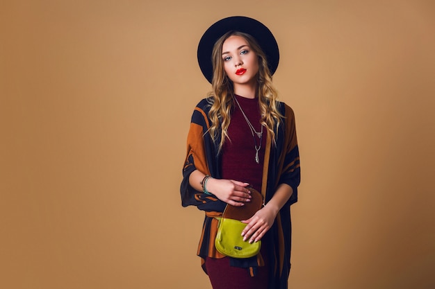 Free photo studio  portrait of young fresh blonde  woman in brown straw poncho , wool black trendy hat and round glasses looking at camera. green leather had bag.