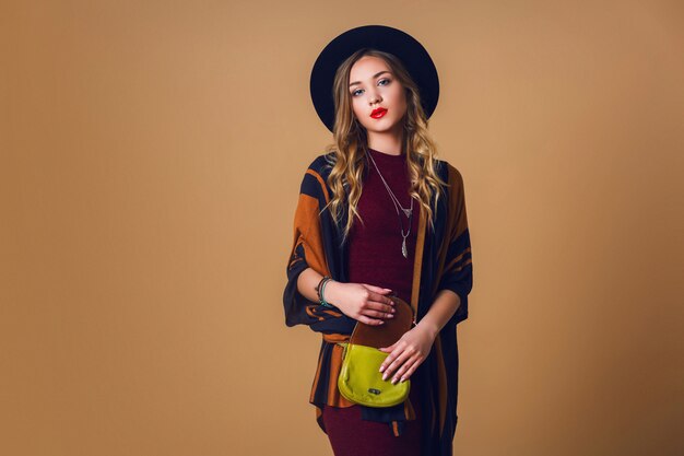 Studio  portrait of young fresh blonde  woman in brown straw poncho , wool black trendy hat and round glasses looking at camera. Green leather had bag.