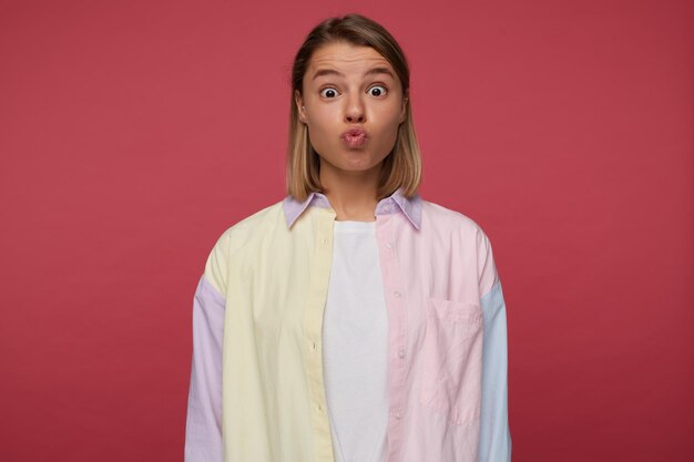 Studio portrait of young funny female posing over red background wears bright shirt shows kiss gesture into camera