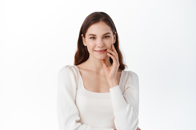 Studio portrait of young woman showing calm and relaxed emotion smiling gently looking at camera touching natural face with nude make up standing against white background