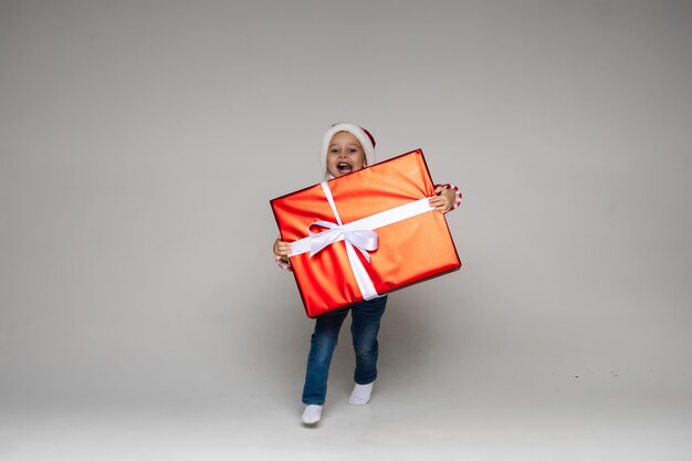 산타 모자와 청바지를 입은 사랑스러운 행복한 어린 소녀의 스튜디오 샷은 흰 활이 달린 빨간 종이에 싸인 큰 크리스마스 선물을 들고 달리고 있습니다.
