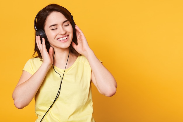 Studio shot of adorable woman with dark hair enjoys of listening to music in headphones