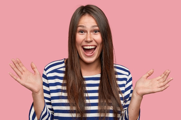 Studio shot of amazed beautiful girl clasps hands with happiness, opens mouth broadly