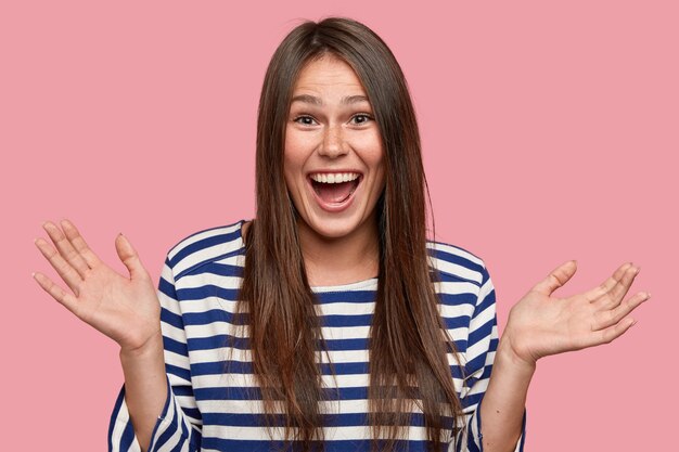 Studio shot of amazed beautiful girl clasps hands with happiness, opens mouth broadly