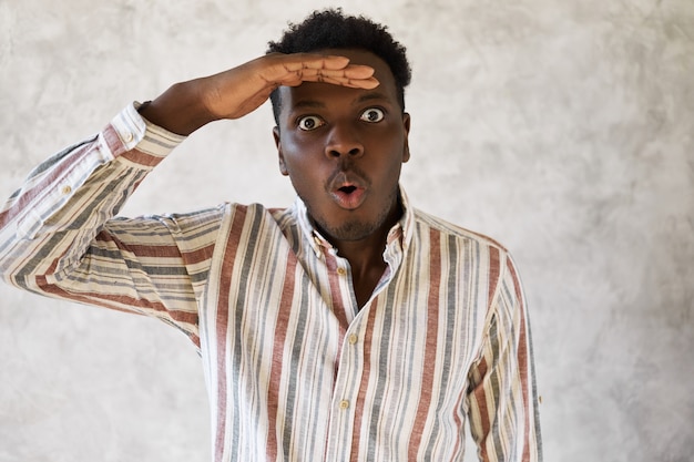 Studio shot of amazed young dark skinned male gaping, keeping hand on forehead, looking into distance, widening eyes because he sees something unexpected.